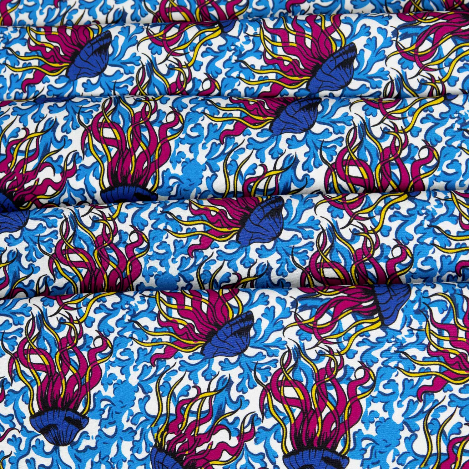 Mussola 100% cotone deadstock con stampa a meduse psichedeliche, tentacoli fucsia e giallo acido su fondo bianco con coralli blu, tessuto piegato in più strati che mostra la consistenza e la morbidezza del cotone leggero estivo